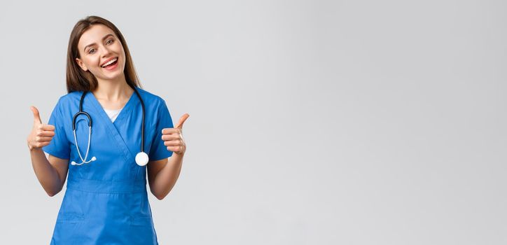 Medical workers, healthcare, covid-19 and vaccination concept. Friendly upbeat female nurse or doctor in blue scrubs, show thumbs-up, stay optimistic, being supportive, assure all good.