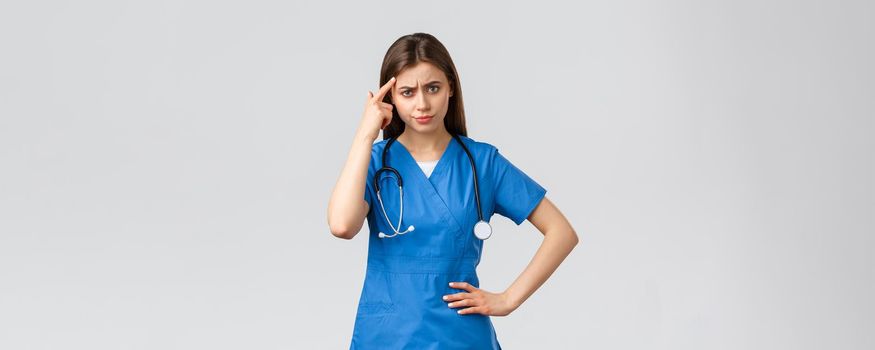 Healthcare workers, prevent virus, insurance and medicine concept. Disappointed angry female nurse, doctor in scrubs pointing at head as scolding patient or coworker, grey background.