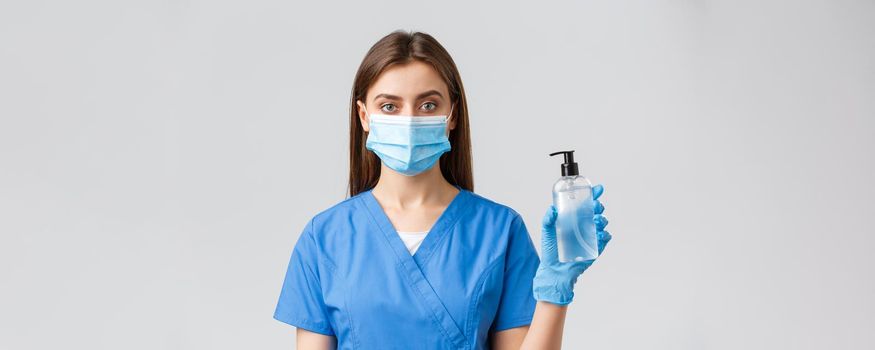 Covid-19, preventing virus, healthcare workers concept. Young pretty professional doctor, female nurse in medical mask and scrubs, showing hand sanitizer or soap to protect from coronavirus.