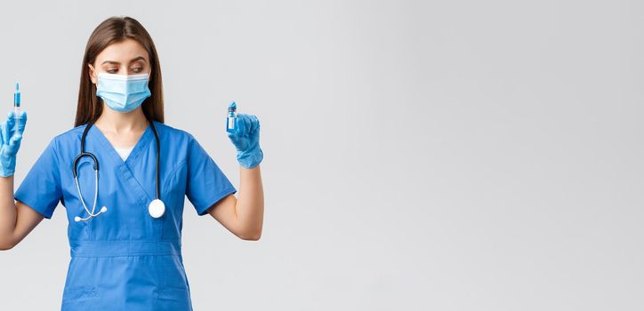 Covid-19, preventing virus, health, healthcare workers and quarantine concept. Female nurse or doctor in blue scrubs, medical mask and gloves, look at ampoule with coronavirus vaccine, hold syringe.