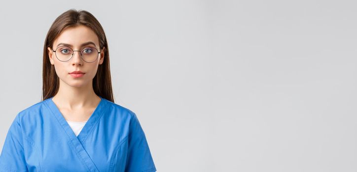 Healthcare workers, medicine, insurance and covid-19 pandemic concept. Young female intern, nurse or doctor in scrubs and glasses, looking at camera with serious expression, grey background.