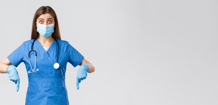 Covid-19, preventing virus, health, healthcare workers and quarantine concept. Excited and interested female doctor, nurse in blue scrubs and medical mask, pointing fingers down and look intrigued.