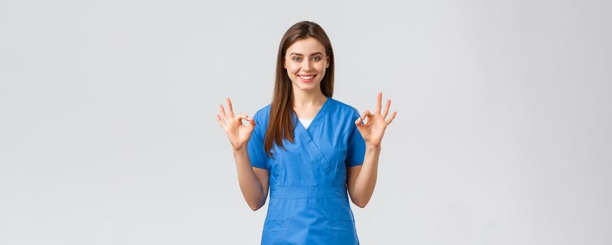 Healthcare workers, prevent virus, insurance and medicine concept. Confident young female nurse, doctor in blue scrubs assure quality of test results in their clinic lub, show okay sign, guarantee.