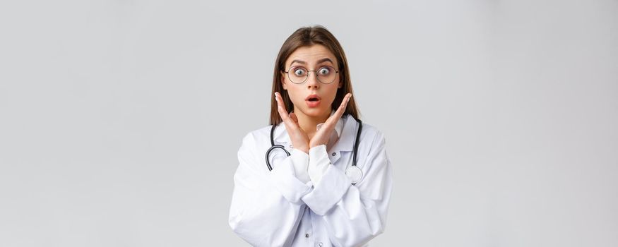 Healthcare workers, medicine, insurance and covid-19 pandemic concept. Surprised and impressed female doctor in glasses, white scrubs, gasping and staring astonished camera, grey background.