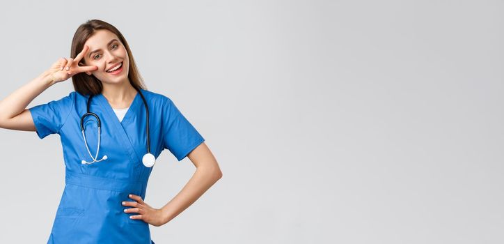 Medical workers, healthcare, covid-19 and vaccination concept. Cheerful attractive female nurse or doctor in blue scrubs, stethoscope, show peace sign, stay optimistic, standing grey background.