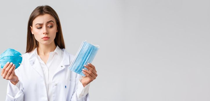 Covid-19, preventing virus, healthcare workers and quarantine concept. Cute female doctor in white scrubs, choosing between medical mask and respirator, pick personal protective equipment.