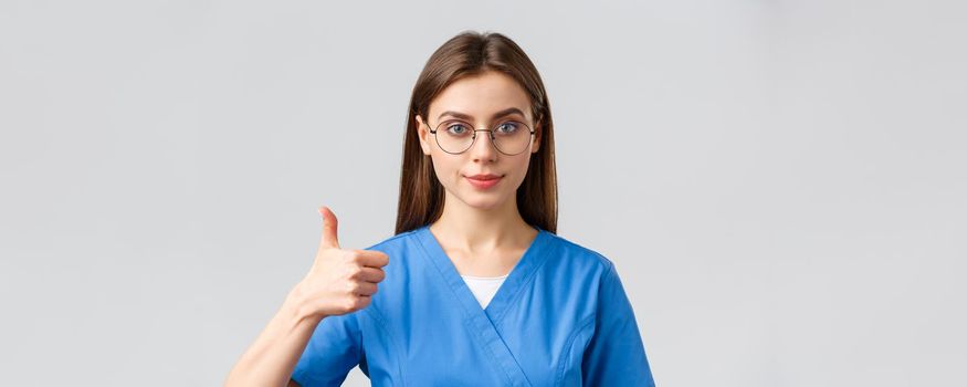 Healthcare workers, medicine, insurance and covid-19 pandemic concept. Close-up supportive and confident female nurse, doctor in blue scrubs and glasses, show approval thumbs-up, guarantee.