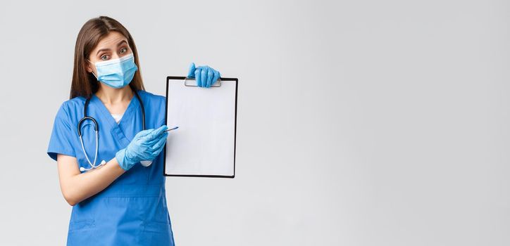 Covid-19, preventing virus, health, healthcare workers and quarantine concept. Friendly female nurse or doctor in blue scrubs and medical mask, explain info on clipboard, pointing paper.