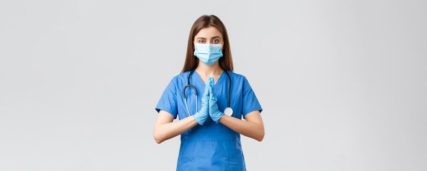 Covid-19, preventing virus, healthcare concept. Pray for medical workers. Female nurse or doctor in blue scrubs, medical mask, fighting coronavirus, press hands together hopeful, pleading.