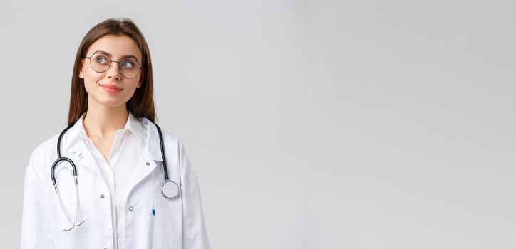 Healthcare workers, medicine, insurance and covid-19 pandemic concept. Dreamy young talented doctor in white scrubs and glasses, looking upper left corner thoughtful and smiling, imaging something.