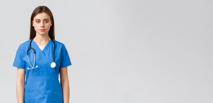 Healthcare workers, prevent virus, insurance and medicine concept. Serious-looking young nurse, doctor in blue scrubs and stethoscope, looking at camera, standing grey background.