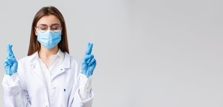 Covid-19, preventing virus, healthcare workers and quarantine concept. Hopeful doctor in white scrubs, medical face mask and gloves, close eyes, cross fingers and praying patients cure infection.