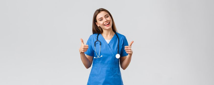 Medical workers, healthcare, covid-19 and vaccination concept. Friendly upbeat female nurse or doctor in blue scrubs, show thumbs-up, stay optimistic, being supportive, assure all good.