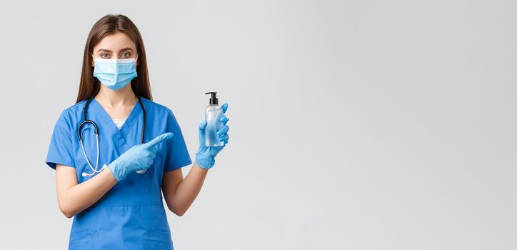 Covid-19, preventing virus, health, healthcare workers and quarantine concept. Young professional female nurse or doctor in blue scrubs, medical mask, pointing finger at hand sanitizer.