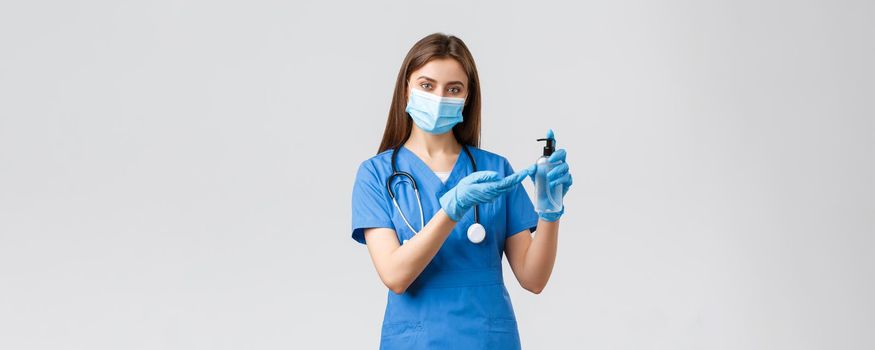 Covid-19, preventing virus, healthcare workers and quarantine concept. Pretty female nurse or doctor in blue scrubs, gloves and medical mask applying hand sanitizer to protect from coronavirus.