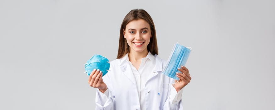 Covid-19, preventing virus, healthcare workers concept. Smiling attractive female doctor in white scrubs showing two variants of personal protective equipment, show respirator and medical mask.