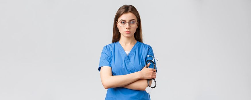 Healthcare workers, medicine, insurance and covid-19 pandemic concept. Serious and determined, professional female nurse, doctor in blue scrubs, holding stethoscope, wear glasses, look confident.