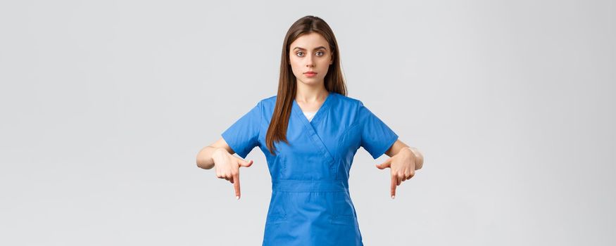Healthcare workers, prevent virus, covid-19 test screening, medicine concept. Serious professional physician, doctor or nurse in blue scrubs, pointing fingers down, provide coronavirus information.