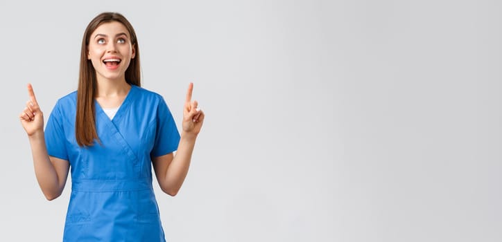 Healthcare workers, prevent virus, covid-19 test screening, medicine concept. Cheerful smiling pretty female doctor or nurse in blue scrubs, pointing fingers up and looking up rejoicing.