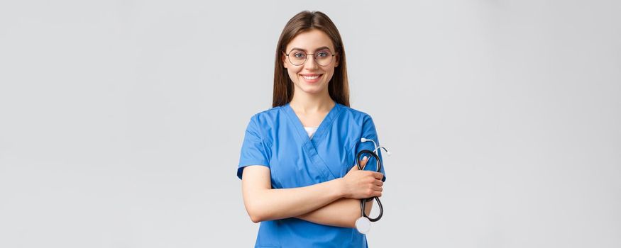 Healthcare workers, medicine, insurance and covid-19 pandemic concept. Confident and enthusiastic young female nurse, doctor in blue scrubs and glasses, hold stethoscope, smiling camera pleased.