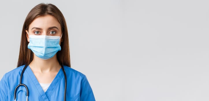 Covid-19, preventing virus, health, healthcare workers and quarantine concept. Close-up young female nurse or doctor in blue scrubs and medical mask looking serious at camera.