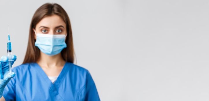 Covid-19, preventing virus, health, healthcare workers and quarantine concept. Serious-looking confident female doctor or nurse in blue scrubs, medical mask, showing syringe with coronavirus vaccine.