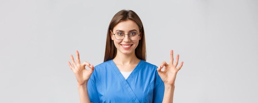 Healthcare workers, medicine, insurance and covid-19 pandemic concept. Confident and optimistic female nurse, doctor in blue scrubs and glasses, show okay sign, guarantee best service quality.