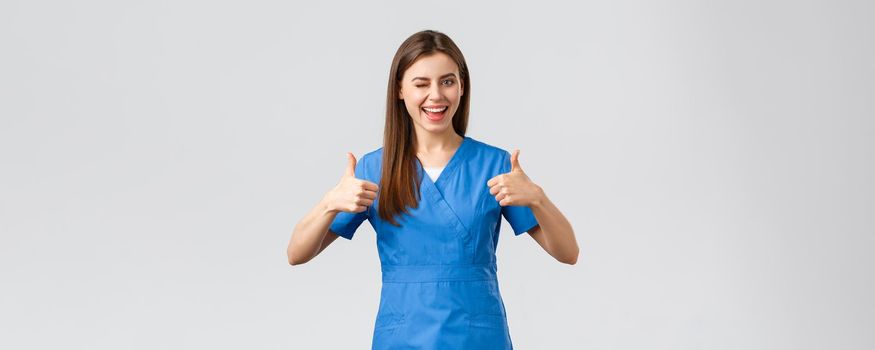Healthcare workers, prevent virus, insurance and medicine concept. Optimistic good-looking female nurse or doctor in scrubs, wink encourage visit clinic, show thumbs-up in approval.