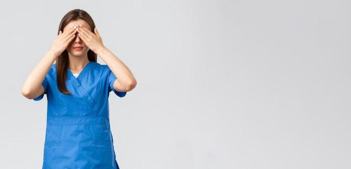 Healthcare workers, prevent virus, insurance and medicine concept. Young woman in blue scrubs, doctor or nurse close eyes with hands, blindfolded medicine, standing grey background.