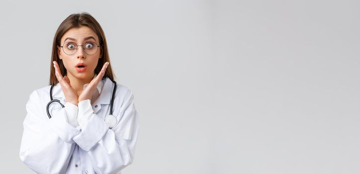 Healthcare workers, medicine, insurance and covid-19 pandemic concept. Surprised and impressed female doctor in glasses, white scrubs, gasping and staring astonished camera, grey background.