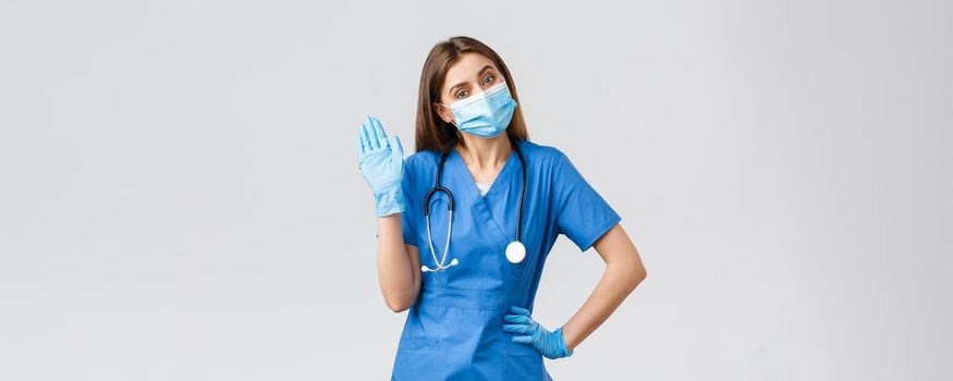 Covid-19, preventing virus, health, healthcare workers and quarantine concept. Friendly female nurse in clinic wearing blue scrubs and medical mask, waving hello or hi sign with gloves on.