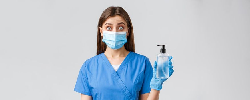 Covid-19, preventing virus, health, healthcare workers and quarantine concept. Excited cute female nurse or doctor in medical mask and scrubs, showing hand sanitizer, look surprised, grey background.