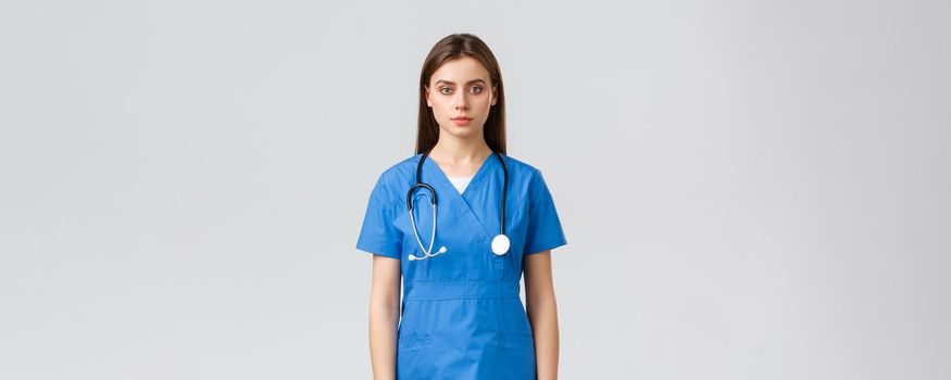 Healthcare workers, prevent virus, insurance and medicine concept. Serious-looking young nurse, doctor in blue scrubs and stethoscope, looking at camera, standing grey background.