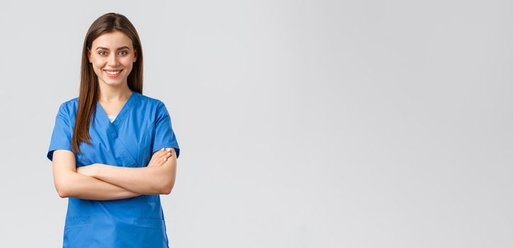 Healthcare workers, prevent virus, insurance and medicine concept. Confident female doctor, nurse in blue scrubs, smiling and cross arms chest, ready to save patient lives, grey background.