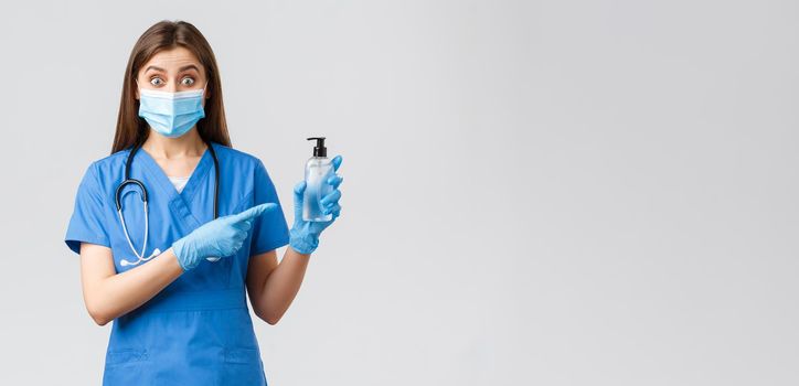 Covid-19, preventing virus, health, healthcare workers and quarantine concept. Enthusiastic female nurse or doctor in blue scrubs, medical mask and gloves, pointing at hand sanitizer, look surprised.