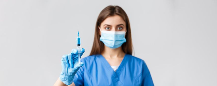 Covid-19, preventing virus, health, healthcare workers and quarantine concept. Serious-looking confident female doctor or nurse in blue scrubs, medical mask, showing syringe with coronavirus vaccine.