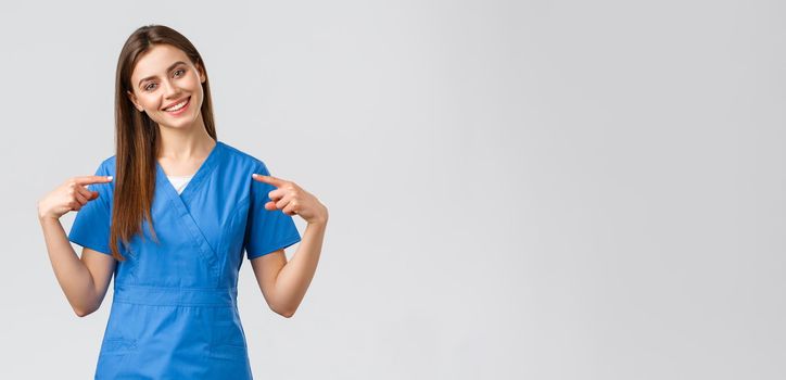 Healthcare workers, prevent virus and medicine concept. Cheerful, smiling pretty doctor, female nurse in blue scrubs pointing at herself, ready to help patients, save lives from coronavirus.