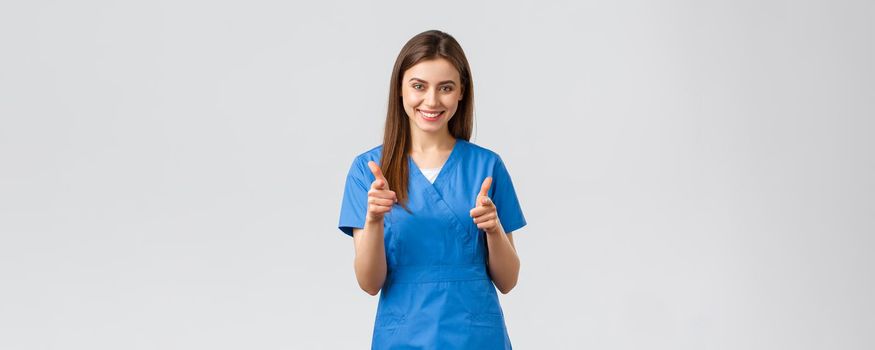Healthcare workers, prevent virus, insurance and medicine concept. Enthusiastic smiling female nurse, doctor in blue scrubs, pointing herself with pistols, choosing, inviting clients to clinic.