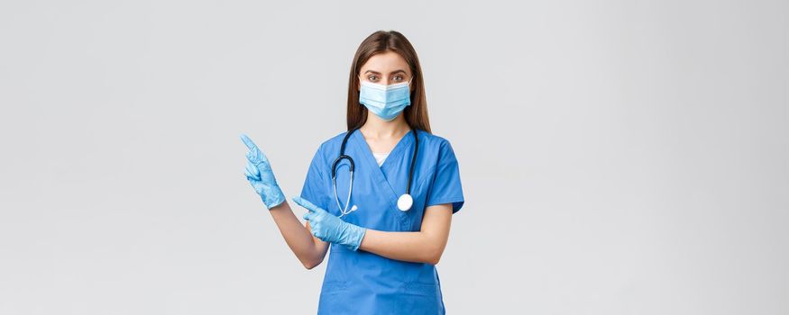 Covid-19, preventing virus, health, healthcare workers and quarantine concept. Pleasant female doctor or nurse in blue scrubs, medical mask and gloves, invite follow page or click link, pointing left.