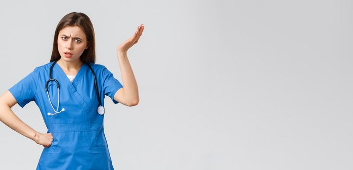 Medical workers, healthcare, covid-19 and vaccination concept. Frustrated and confused young female nurse, pretty doctor in blue scrubs, hear strange idea, shrug with hand raised in nonsense.