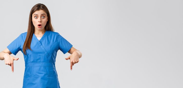 Healthcare workers, prevent virus, covid-19 test screening, medicine concept. Shocked and surprised female nurse or doctor in blue scrubs, pointing fingers down to inform people about promo.