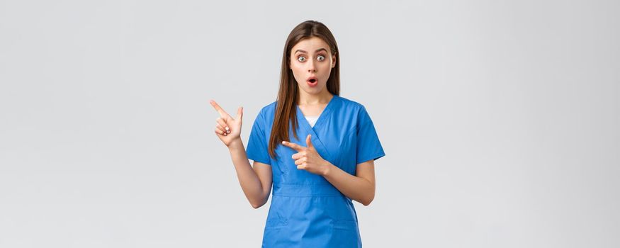 Healthcare workers, prevent virus, insurance and medicine concept. Surprised impressed nurse or doctor in blue scrubs, open mouth and stare camera, pointing fingers left astonished.