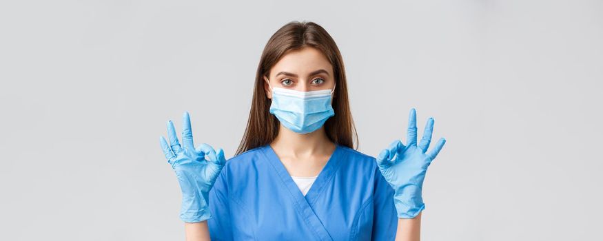 Covid-19, preventing virus, health, healthcare workers and quarantine concept. Professional female nurse or doctor in clinic, blue scrubs and medical mask, show okay sign, all under control.