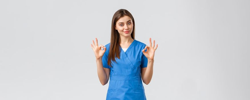 Healthcare workers, prevent virus and medicine concept. Attractive confident female nurse, doctor in blue scrubs, smirk show okay sign, guarantee quality of service in clinic, recommend medicine.
