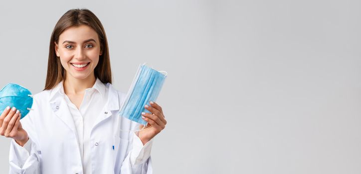 Covid-19, preventing virus, healthcare workers concept. Smiling attractive female doctor in white scrubs showing two variants of personal protective equipment, show respirator and medical mask.