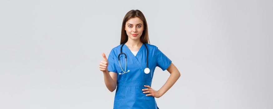 Medical workers, covid-19 and vaccination concept. Confident professional doctor, female nurse in blue scrubs and stethoscope, show thumbs-up, assure or guarantee best quality of service in clinic.