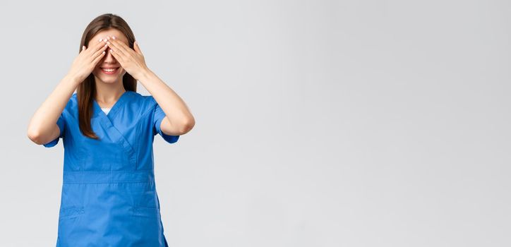 Healthcare workers, prevent virus, insurance and medicine concept. Young optimistic female nurse, doctor in scrubs, shut eyes with hands and smiling, anticipating surprise, being blindfolded.