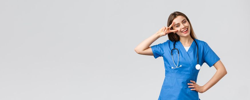 Medical workers, healthcare, covid-19 and vaccination concept. Cheerful attractive female nurse or doctor in blue scrubs, stethoscope, show peace sign, stay optimistic, standing grey background.