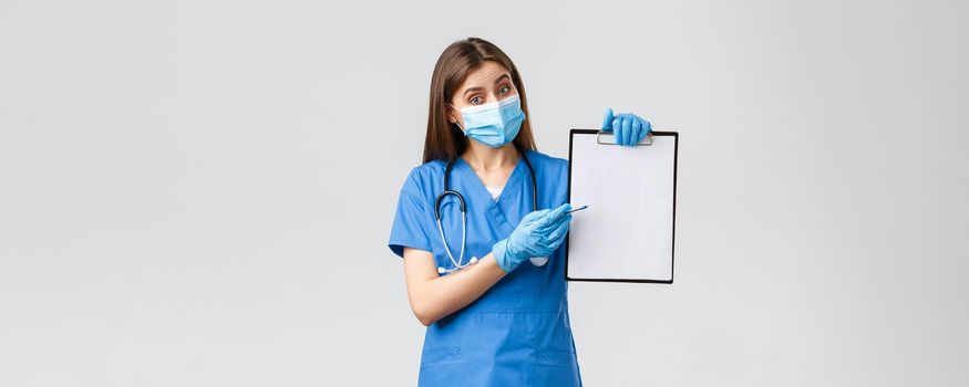 Covid-19, preventing virus, health, healthcare workers and quarantine concept. Friendly female nurse or doctor in blue scrubs and medical mask, explain info on clipboard, pointing paper.