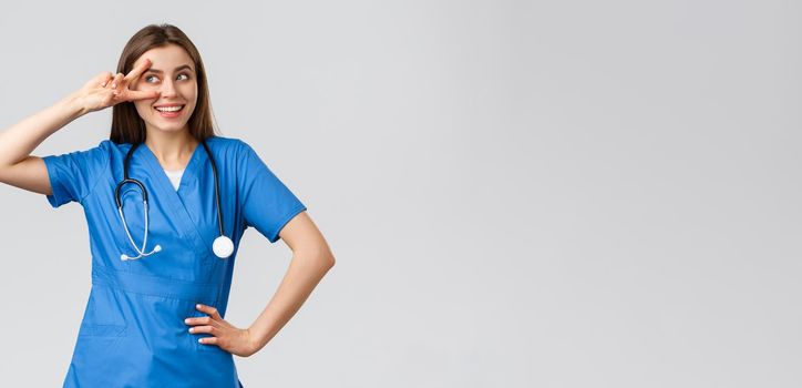 Medical workers, healthcare, covid-19 and vaccination concept. Cheerful and energized young female doctor, pretty nurse in blue scrubs, stethoscope, show peace near eye and smiling upbeat.